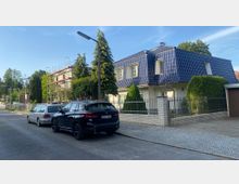 Straßenszene in einem Wohngebiet mit einem Einfamilienhaus mit blauer Dachfläche und hellem Putz, umgeben von einem Zaun und gepflegtem Vorgarten. Im Hintergrund ein mehrstöckiges Gebäude mit Gerüst sowie geparkte Autos entlang der Straße.