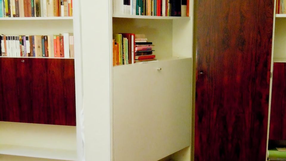 Innenraum mit eingebauten Bücherregalen aus weißem Holz und dunkel furnierten Schranktüren; ein schwenkbares Regal ist geöffnet und zeigt mehrere Reihen mit Büchern. Holzboden und eine niedrige Decke rahmen die Regalanlage ein.
