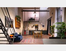 Offener Wohnraum im Industrial-Stil mit hohen Decken, einer Metalltreppe zu einer kleinen Zwischenterrasse, einer offenen Küche mit Holztisch und Stühlen sowie mehreren Pflanzen. Große Fenster an der rechten Seite lassen Tageslicht herein, ergänzt durch Wandleuchten und hängende Lampen.