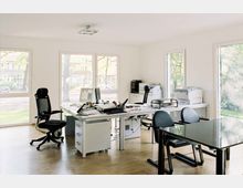 Modernes Büro mit hellen Wänden und großen Fensterfronten, die Blick auf eine grüne Außenanlage bieten. Der Raum enthält mehrere Schreibtische mit Bürostühlen, Computern, Druckern sowie Aktenstapeln, und ein zusätzlicher Besprechungstisch mit schwarzen Stühlen.