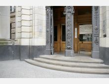 Eingangsbereich eines historischen Gebäudes mit steinernen Säulen und kunstvoll verzierten Metallpfosten neben einer doppelflügeligen Holztür. Im Vordergrund befinden sich halbrunde Stufen aus Stein, die zum Eingang führen.