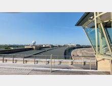 Blick auf das Dach des ehemaligen Flughafens Tempelhof in Berlin mit flachen Gebäudestrukturen, einer großen Rollbahn im Hintergrund und einer Beobachtungsplattform mit Glasfront im Vordergrund. Am Horizont ist die Skyline der Stadt bei klarer Sicht zu erkennen.