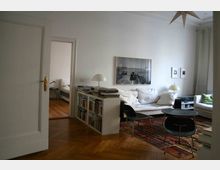 Helles Wohnzimmer mit Holzboden, einem weißen Sofa, einem niedrigen schwarzen Couchtisch, einem Bücherregal mit Büchern und Schreibtischlampen sowie einem bunten Teppich. Im Hintergrund führt eine offene Tür in ein Schlafzimmer mit einem Bett.