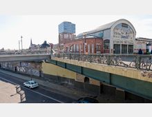Eine städtische Szene zeigt eine Fußgängerbrücke mit angeketteten Fahrrädern, darunter eine Straße mit geparkten Autos und Graffiti an den Wänden. Im Hintergrund befinden sich ein historisches Backsteingebäude mit großen Glasfenstern und Rundbogen sowie ein moderner Büroturm.