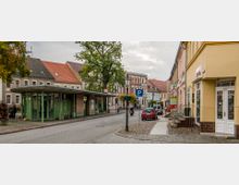 Straßenszene in einer kleinen Stadt mit Kopfsteinpflaster, Wohnhäusern und Geschäften im klassischen Stil. Links im Vordergrund befindet sich eine Bushaltestelle mit einem grünen Überdach, während rechts pastellfarbene Gebäude und ein geparktes rotes Auto zu sehen sind.