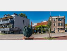 Moderne Wohnhäuser und ein Backsteingebäude stehen um eine kleine gepflasterte Fläche, in deren Mitte eine bronzene Skulptur mit Figuren in einem Boot platziert ist. Im Hintergrund sind Dächer mit roten Ziegeln und Grünpflanzen zu sehen, unter einem klaren blauen Himmel.