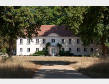 Zweigeschossiges, helles Herrenhaus mit symmetrischen Fenstern, einem roten Ziegeldach und einem kleinen Balkon über dem Haupteingang. Der Weg zum Gebäude ist von Rasenflächen und hohen Bäumen gesäumt.