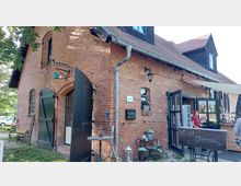 Backsteinhaus mit roten Ziegeln, grün gestrichenen Holztüren und einem Schilderhalter in Form einer Waage. Auf der rechten Seite befindet sich ein Eingangsbereich mit Glastüren, darüber ein Vordach. Dekorationen wie Blumentöpfe und Laternen schmücken den Eingangsbereich, während eine Holztafel mit der Aufschrift „Herzlich Willkommen“ vor dem Eingang steht.