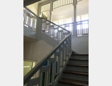 Treppenhaus in einem historischen Gebäude mit Holzstufen und einem verzierten weißen Geländer. Große Fenster im oberen Bereich lassen Tageslicht in den Raum.