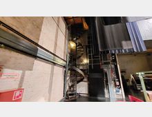 Bühnenbereich mit freiliegender Backsteinwand, einer Wendeltreppe aus Metall und hängenden schwarzen Vorhängen. Im Vordergrund sind technische Einrichtungen und ein Schild mit der Aufschrift „Löschwasserstelle freihalten“ zu erkennen.