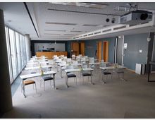 Seminarraum mit grauen Wänden, rechteckigen Tischen und gepolsterten Stühlen. Der Raum verfügt über große Fenster auf der linken Seite, eine Deckenbeleuchtung, Projektor und eine Rednerbühne im vorderen Bereich.