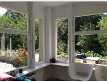 Blick aus einem hellen Raum mit großen weißen Fenstern auf eine Terrasse und einen Garten mit Bäumen und Sträuchern. Auf der Terrasse stehen Blumentöpfe, ein Grill, und dahinter breitet sich eine grüne, bewaldete Umgebung aus.