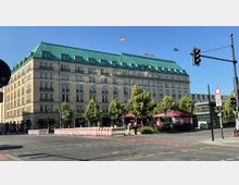 Straßenszene mit dem Gebäude des Hotels Adlon in Berlin, einem mehrstöckigen, historischen Gebäude mit hellem Fassadenputz und grünem Mansardendach. Im Vordergrund sind Bäume, ein Gehweg mit roten Sonnenschirmen und eine Straßenkreuzung mit Ampeln zu sehen.