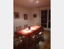 Esszimmer mit Holzboden und einem rechteckigen Esstisch, der mit einer rot-orange karierten Tischdecke gedeckt ist. Der Tisch ist von vier Holzstühlen umgeben, und ein blaues Glasgefäß steht darauf. Im Hintergrund sind eine weiße Wand mit Bildern und eine verglaste Terrassentür mit Vorhängen zu sehen.