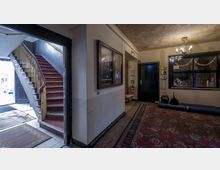 Flur eines alten Gebäudes mit roten Teppichen, einer offenen Holztreppe mit gelbem Geländer links und dunklen Türen. An der Wand hängen eine Glasvitrine sowie ein Wandtelefon, und die Decke zeigt sichtbare Gebrauchsspuren.