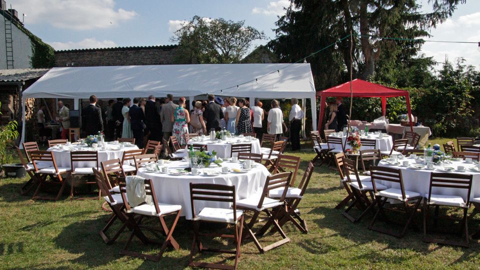 Gartenbereich mit runden, weiß gedeckten Tischen und Holzklappstühlen auf einer Rasenfläche, daneben ein großes weißes Zelt und ein rotes Pavillonzelt. Im Hintergrund stehen hohe Bäume, Büsche und eine niedrige Mauer bzw. Gebäudefassade.