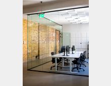 Glasverkleideter Besprechungsraum in einem modernen Büro mit freiliegender Ziegelwand, langem weißen Tisch und mehreren Schreibtischstühlen mit Monitoren. An der Decke sind rechteckige Lichtpaneele und ein beleuchtetes Notausgangsschild sichtbar.