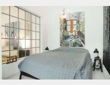 Minimalist bedroom with white walls, a double bed covered in a blue patterned quilt, and black bedside lamps on small tables. A large frosted grid-style window and a colorful abstract painting above the headboard define the space.