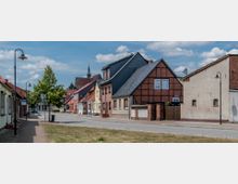 Straßenszene in einer kleinen Stadt mit Kopfsteinpflaster, rechts Fachwerkhäuser und verputzte Gebäude mit Satteldächern. Im Hintergrund ein Kirchturm, links Gehwege mit Laternen und Bäumen.