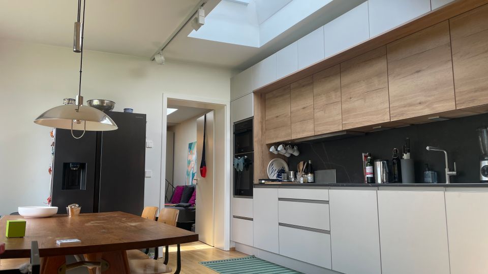 Moderne Wohnküche mit langen weißen und holzfarbenen Einbauschränken, dunkler Arbeitsfläche und Spüle sowie schwarzem Kühlschrank neben einem Esstisch aus Holz. Ein großes Oberlicht im Dach und Schienenstrahler sorgen für viel Tageslicht, im Hintergrund führt eine Türöffnung in einen angrenzenden Raum.