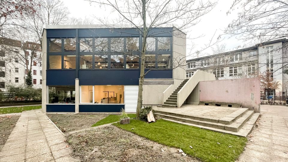 Zweistöckiges modernes Gebäude mit dunkelblauer Fensterfassade und großen Glasflächen, gelegen in einem Innenhof mit Rasenflächen und kahlen Bäumen. Rechts führt eine breite Außentreppe aus Beton zu einer erhöhten Plattform neben einer niedrigen Mauer.