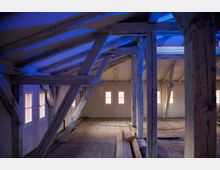 Dachboden eines historischen Gebäudes mit sichtbaren, weiß gestrichenen Holzbalken und Holzboden. Mehrere kleine Fenster lassen Licht in den Raum, der von blauen Lichtreflexen beleuchtet wird.