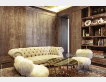 Heller Wohnraum mit einer beigen, gepolsterten Chesterfield-Sitzbank, einem runden Marmortisch mit goldenem Gestell und zwei Sesseln mit flauschiger Polsterung. Im Hintergrund befindet sich eine dunkle Holzvertäfelung sowie ein Bücherregal mit Büchern und dekorativen Objekten.