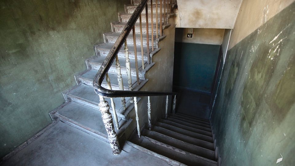 Schmales Treppenhaus in einem älteren Gebäude mit abgenutzten Holzstufen und einem schwarzen Handlauf mit weißen, abgeplatzten Geländerstäben. Die Wände sind zweifarbig grün und beige gestrichen und zeigen starke Gebrauchsspuren sowie abgeblätterte Farbe.