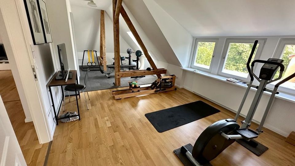 Dachgeschossraum mit Holzboden und Fitnessgeräten, darunter ein Crosstrainer, eine Hantelbank und ein Rudergerät; sichtbare Dachschrägen, freiliegende Holzbalken und große Fenster, die viel Tageslicht hereinlassen.