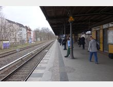 Ein Bahnhofssteig mit überdachter Wartezone und gelben Wandfliesen, an dem mehrere Personen stehen. Rechts befinden sich Fahrpläne und Türen, während links die Gleise verlaufen, flankiert von Bäumen und Wohnhäusern.