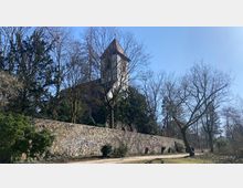 Kirche mit spitzem Turmdach auf einem Hügel, umgeben von Bäumen. Im Vordergrund eine Steinmauer und ein Gehweg, teilweise von Sträuchern und kahlen Bäumen überschattet.