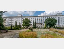 Großes, weißes mehrstöckiges Gebäude im klassischen Stil mit regelmäßigen Fenstern und dekorativen Details, umgeben von einem gepflegten Garten. Im Vordergrund befindet sich ein kleiner Teich mit Schilfpflanzen und einer Wasserfontäne, sowie ein Weg mit einer Holzbank.