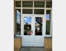 Weiße Doppeltür mit Glaseinsätzen in einem Gebäude mit gelber Klinkerfassade, durch die man eine Außentreppe und umliegende Gebäude bei Tageslicht sieht. Über der Tür befinden sich zusätzliche Glasfenster, die den Eingangsbereich öffnen.