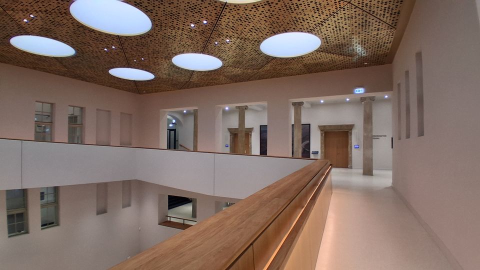 Innenansicht eines modernen Atriums mit umlaufender Galerie und hellem Geländer mit Holzhandlauf. Die Decke ist mit perforierten Paneelen und großen ovalen Oberlichtern gestaltet; entlang der Wände stehen Säulen und mehrere Türöffnungen.