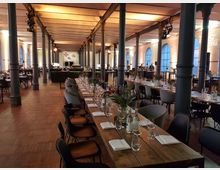 Großer Veranstaltungsraum mit langen, gedeckten Holztischen, umgeben von schwarzen Pfeilern. Die Halle hat hohe Decken, warme Beleuchtung und große Fenster mit blauen Rahmen an den Wänden.