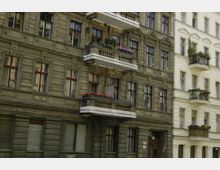 Fassade eines Altbaus mit dekorativen Verzierungen und Balkonen, teils mit Blumen geschmückt. Die linke Gebäudeseite ist dunkel und verwittert, während die rechte Seite heller und renovierter wirkt.