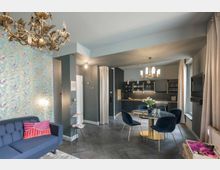 Modern eingerichteter Wohn- und Essbereich in einer Wohnung mit dunkelgrauen Wänden, einem Sofa, einer floral gemusterten Tapete, einer kleinen Küche mit schwarzen Schränken und goldenen Akzenten sowie einem runden Esstisch mit blauen Polsterstühlen. Ein dekorativer Kronleuchter und Fenster mit Vorhängen sorgen für zusätzliche Details.