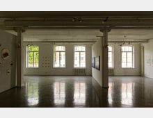 Industrie-inspirierter Ausstellungsraum mit glänzendem Betonboden, weißen Wänden und großer Fensterfront aus rechteckigen Fenstern. An den Wänden hängen Kunstwerke, und sichtbare Deckenrohre betonen das minimalistische Design.