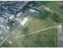 Luftaufnahme eines kleinen Flughafens mit Start- und Landebahn, mehrere kleine Gebäude und Hangars am Rand. Umgeben von landwirtschaftlichen Flächen und Gebäuden eines angrenzenden Stadtteils.