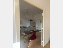 Blick in eine modern eingerichtete Küche mit weißer Einbauzeile, darüber Hängeschränke und einer freistehenden Kühl-Gefrierkombination. Im Vordergrund ein roter Esstisch mit Blumen und Obstschale, daneben Holz- und Polsterstühle, sowie Parkettboden und eine verzierte Deckenleuchte.