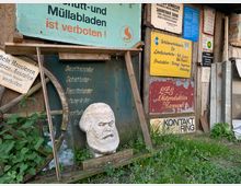 Außenansicht einer alten Holzwand mit mehreren verwitterten Schildern, darunter Werbe- und Verbotsschilder sowie eine steinerne Karl-Marx-Büste im Vordergrund. Daneben sind ein altes hölzernes Wagenrad und Pflanzen zu sehen, die am Boden wachsen.