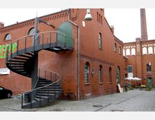Roter Backsteinbau mit rundbogigen Fenstern und einem außen angebrachten, schwarzen Wendeltreppenaufgang, der zu einer grünen Tür im ersten Stock führt. Der gepflasterte Innenhof ist teils mit Fahrrädern, Schildern und Sonnenschirmen ausgestattet; im Hintergrund ist ein hoher Schornstein zu sehen.