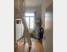 Kinderzimmer mit Hochbett aus Holz und Leiter, hellem Holzfußboden, einem großen Fenster mit dünnem Vorhang und Spielzeug sowie Aufbewahrungseinheiten entlang der Wände. Ein runder Teppich liegt unter dem Bettbereich.