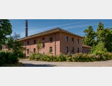 Zweistöckiges rosa Gebäude mit Satteldach, umgeben von grünen Bäumen und blühenden Büschen. Im Hintergrund ist ein hoher Industrieschornstein zu sehen, und der Himmel ist klar.