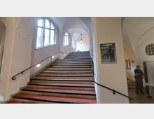 Breiter Treppenaufgang in einem historischen Gebäude mit cremefarbenen Wänden, großen Rundbogenfenstern und einer Deckenleuchte. Die Treppe ist auf beiden Seiten mit Handläufen versehen, und an der Wand hängt ein Informationsplakat.