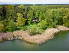 Haus im A-Frame-Stil, umgeben von dichter Vegetation, am Ufer eines Sees mit Schilfbewuchs. Im Hintergrund erstreckt sich ein bewaldetes Gebiet.