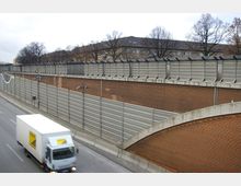 Straßenabschnitt mit Schallschutzwänden aus Metall und Backstein-Mauerwerk, darunter eine geteerte Fahrbahn mit einem fahrenden weißen Lkw. Im Hintergrund sind mehrstöckige Wohngebäude und kahle Bäume sichtbar.