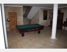 Ein Innenraum mit einem Billardtisch in der Mitte, beige gefliesten Böden und einer Treppe mit Metallgeländer im Hintergrund. An der Wand befinden sich eine Holztür und Dekorationen, während an den Seiten antike Holzmöbel und eine weiße Säule stehen.