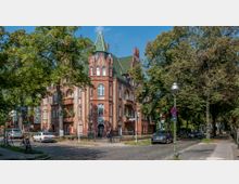 Eckgebäude im Stil der Backsteingotik mit grünem Spitzdach und verzierten Fenstern, umgeben von Bäumen und parkenden Autos auf einer gepflasterten Straße. Im Vordergrund sind Laternen und Straßenschilder zu sehen, mit einer ruhigen Wohngegend im Hintergrund.