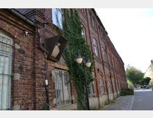 Backsteinfassade eines historischen Gebäudes mit hohen Fenstern, teilweise von Efeu bewachsen, und Gitterstäben vor den unteren Fenstern; im Vordergrund eine zweiflammige Straßenlaterne an einem gepflasterten Gehweg.